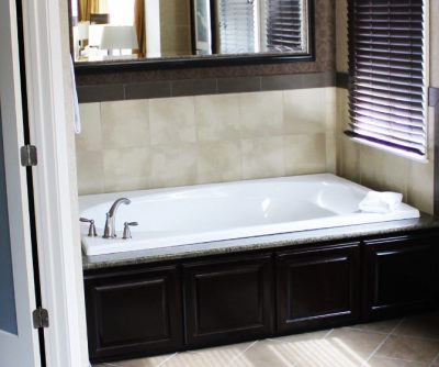 Spacious hotel bathroom featuring a large white soaking tub, granite countertop, and natural light from window blinds.