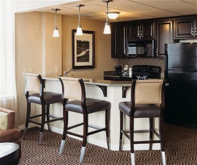 Modern hotel kitchenette with dark wood cabinetry, counter seating for three, and appliances including a refrigerator and mic